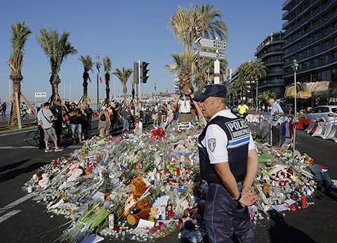 A police officer watches people gathering around a floral tribute for the victims killed during a deadly attack, on the famed Boulevard des Anglais in Nice, southern France, Sunday, July 17, 2016. French authorities detained two more people Sunday in the investigation into the Bastille Day truck attack on the Mediterranean city of Nice that killed at least 84 people, as authorities try to determine whether the slain attacker was a committed religious extremist or just a very angry man. (AP Photo/Laurent Cipriani)