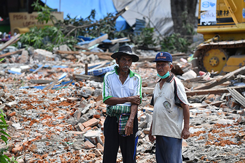 Two residents look on as damaged houses are being destroyed by workers using an excavator after an earthquake hit the area of Gangga on August 12, 2018. - An earthquake on the Indonesian island of Lombok has killed 387 people, authorities said on August 11, adding hundreds of thousands of displaced people were still short of clean water, food and medicine nearly a week on. (Photo by ADEK BERRY / AFP)