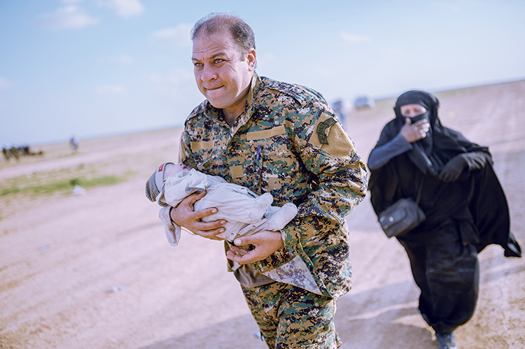 Mostapha Bali, the spokesman for the Kurdish-led Syrian Democratic Forces (SDF), carries a baby followed by a woman after fleeing the Islamic State (IS) group's last holdout of Baghouz, in Syria's northern Deir Ezzor province, on February 22, 2019. (Photo by Bulent KILIC / AFP)