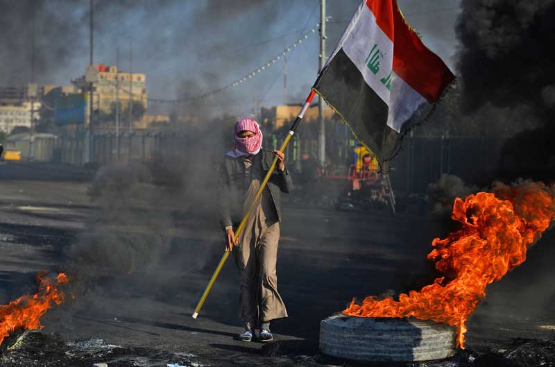 TOPSHOT - CORRECTION - An anti-government protester waves a national flag as he stands before flaming tires at a make-shift roadblock during a demonstration in the central Iraqi holy shrine city of Najaf on January 20, 2020. (Photo by Haidar HAMDANI / AFP) / The erroneous mention[s] appearing in the metadata of this photo by Haidar HAMDANI has been modified in AFP systems in the following manner: [January 20] instead of [January 19]. Please immediately remove the erroneous mention[s] from all your online services and delete it (them) from your servers. If you have been authorized by AFP to distribute it (them) to third parties, please ensure that the same actions are carried out by them. Failure to promptly comply with these instructions will entail liability on your part for any continued or post notification usage. Therefore we thank you very much for all your attention and prompt action. We are sorry for the inconvenience this notification may cause and remain at your disposal for any further information you may require.