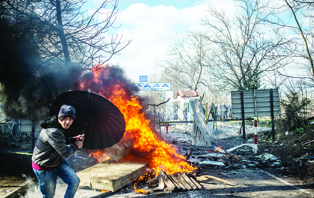 TOPSHOT - A man takes coover behind an umbrella as he throws a mattress in a fire during clashes with Greek police in the buffer zone at Turkey-Greece border, at Pazarkule, in Edirne district, on February 29, 2020. - Thousands of migrants stuck on the Turkey-Greece border clashed with Greek police on February 29, 2020, according to an AFP photographer at the scene. Greek police fired tear gas at migrants who have amassed at a border crossing in the western Turkish province of Edirne, some of whom responded by hurling stones at the officers. The clashes come as Greece bolsters its border after Ankara said it would no longer prevent refugees from crossing into Europe following the death of 33 Turkish troops in northern Syria. (Photo by BULENT KILIC / AFP)
