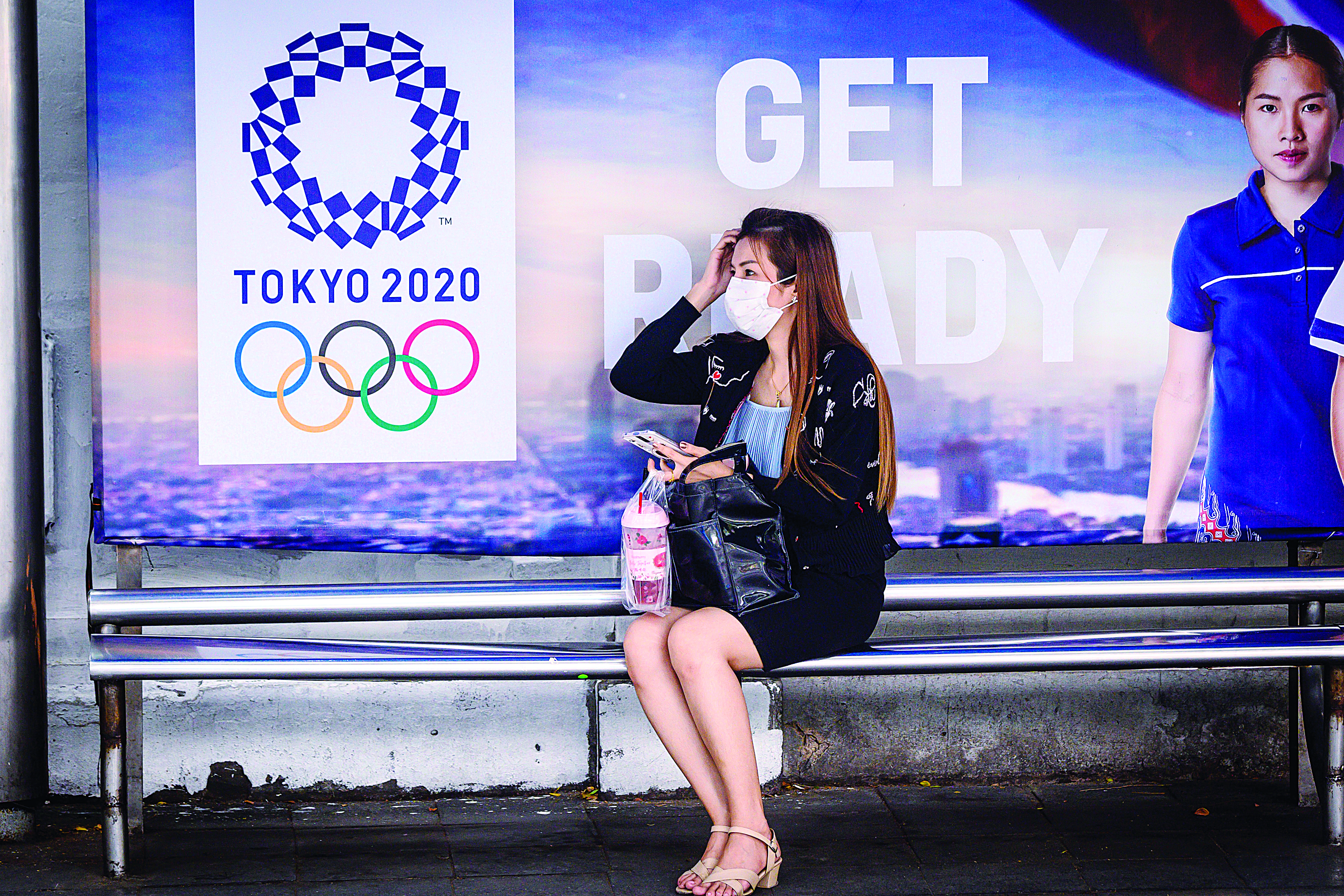 A woman wearing a facemask, amid concerns over the spread of the COVID-19 coronavirus, sits at a bus stop in front of a Tokyo 2020 Olympics advertisement in Bangkok on March 16, 2020. (Photo by Mladen ANTONOV / AFP)