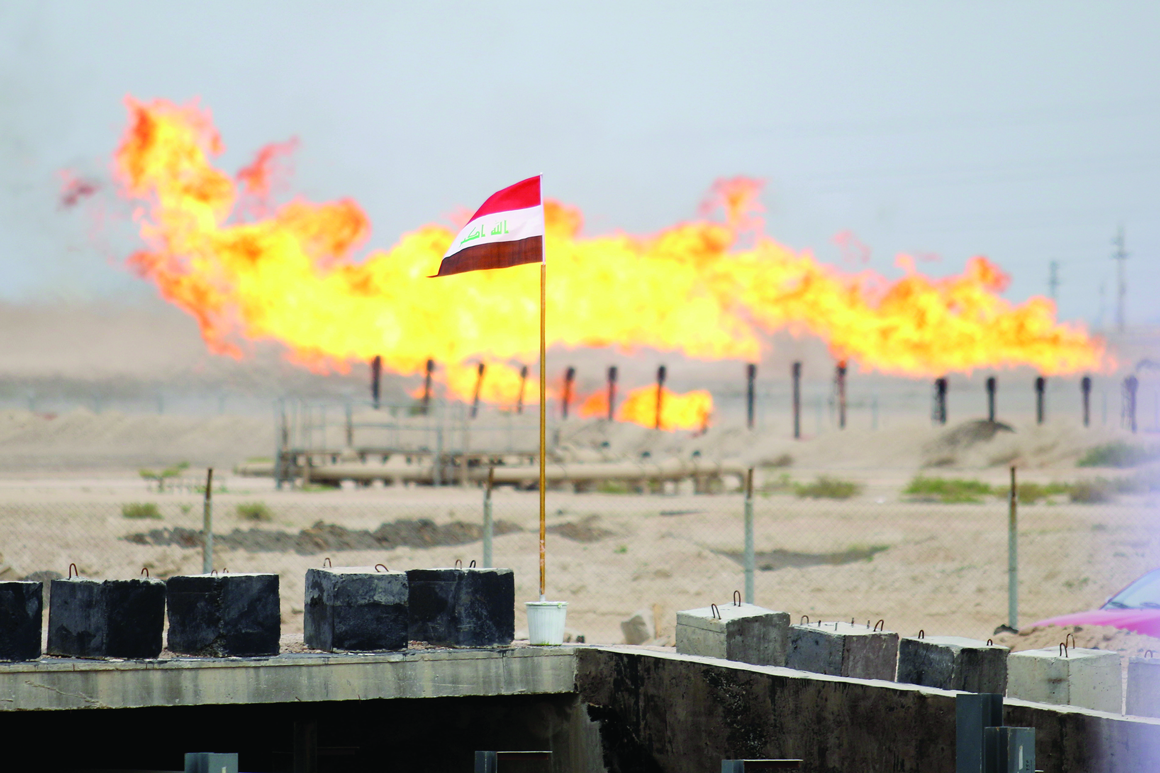 (FILES) This file photo taken on March 3, 2016, shows a an Iraqi national flag flying before front of excess gas being burnt off, at a pipeline in the newly opened section of the oil refinery of Zubair, southwest of Basra in southern Iraq. - As crude prices plunge, Iraq's oil sector is facing a triple threat that has slashed revenues, risks denting production and may spell trouble for future exports. (Photo by HAIDAR MOHAMMED ALI / AFP)