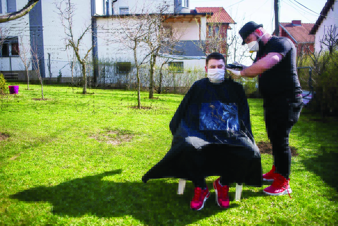Kosovo hairdresser Driton Kameri (R) wears a facemask as he cuts the hair of a client in his garden on March 30, 2020 in Pristina during the time of the COVID-19 pandemic. - In Pristina, a barber is defying coronavirus edits, such as the ban on opening non-essential shops, to practice his art in the open air, but not without first spraying his customers with disinfectant from head to toe. (Photo by Armend NIMANI / AFP)