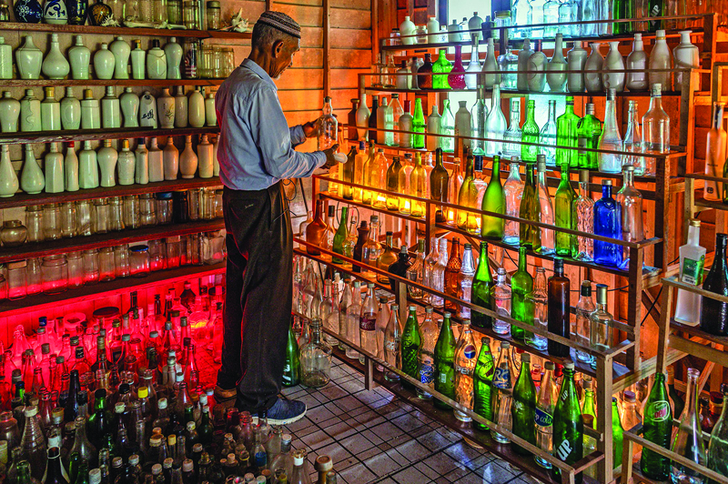 TOPSHOT - This picture taken on September 12, 2020 shows Tengku Mohamad Ali Mansor inspecting a glass bottle at his bottle museum in Penarik village in the Setiu district of the eastern Malaysian state of Terengganu. - An elderly Malaysian's quest to rid the country's beaches of washed-up glass led to him amassing a collection of thousands of bottles, now displayed in a colourful seaside museum. (Photo by Mohd RASFAN / AFP)