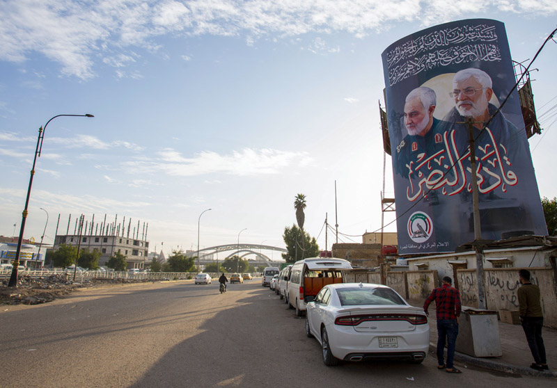 Iraqi men stand beneath a billboard depicting late top Iranian commander Qasem Soleimani (L) and leading Iraqi paramilitary figure Abu Mahdi al-Muhandis, on a street in the southern city of Basra, on December 28, 2020, on the eve of the one-year anniversary of their killing in a US drone strike in the Iraqi capital in January. (Photo by Hussein FALEH / AFP)