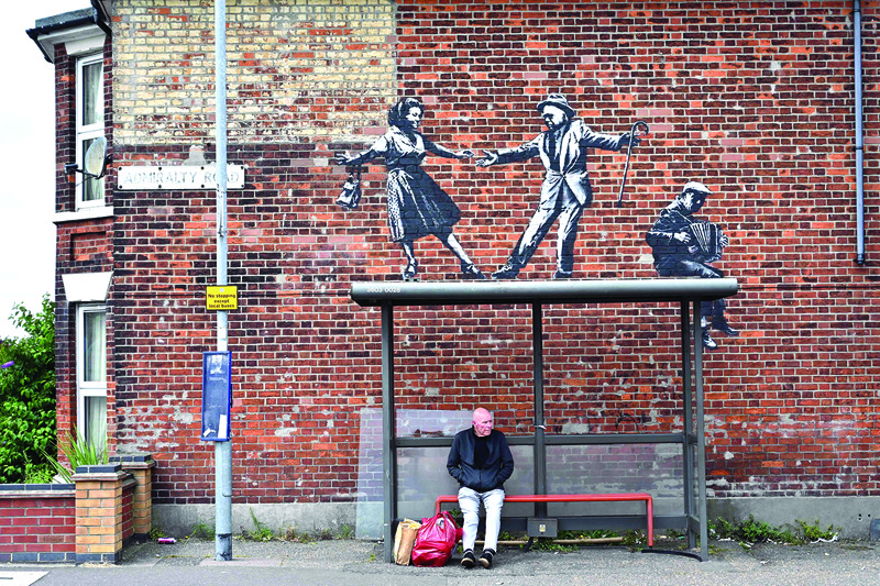 A man waits at a bus stop below a Bansky graffiti artwork of a couple dancing to an accordion player on a wall in Great Yarmouth on the east coast of England.n