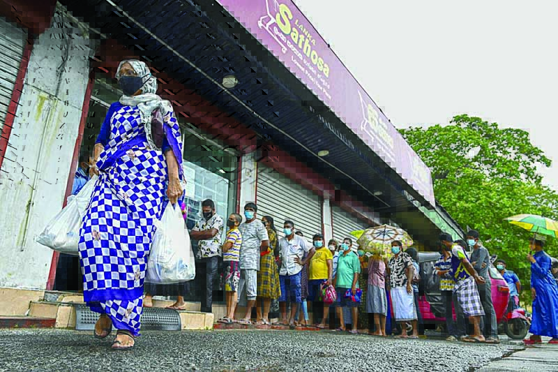 COLOMBO: A woman carrying food bags walks pasts people standing in queue outside a state-run supermarket to buy essential food items in Colombo Friday as Sri Lanka began imposing price controls on essential food from September 3 after using a state of emergency to seize allegedly hoarded stocks of sugar and rice. - AFPn