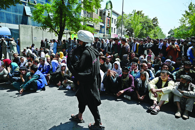 KABUL: A Taleban fighter walks in front of people sitting along a road outside a bank waiting to withdraw money in the Shar-e-Naw neighborhood yesterday. - AFP  n