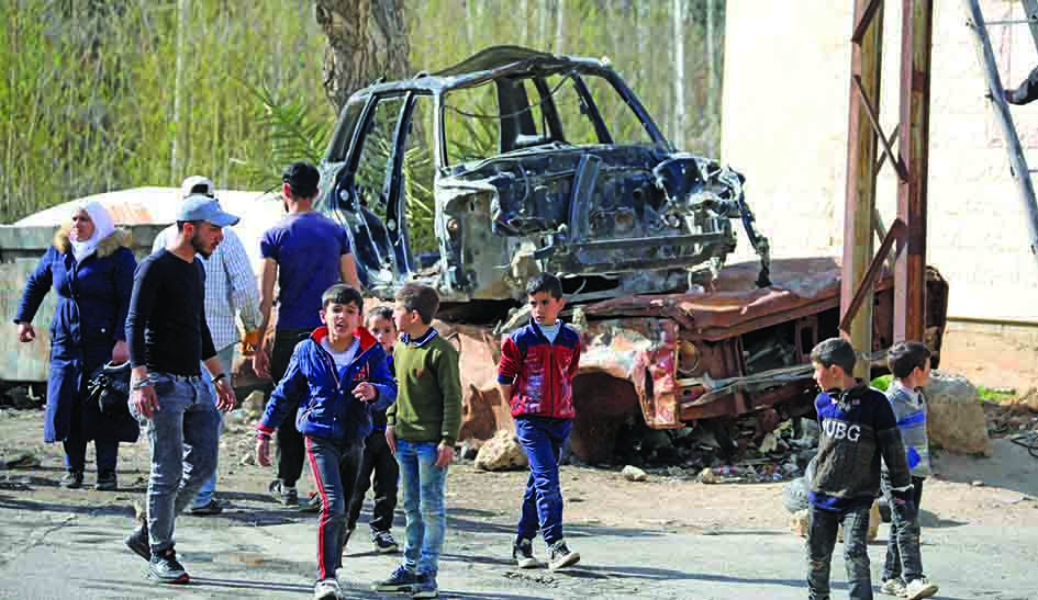 Children walk by the carcass of a burnt car during the filming of the Syrian Social series 