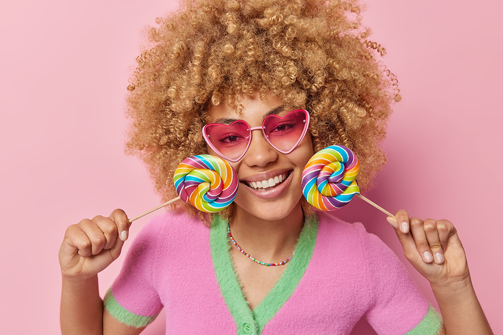 Indoor shot of happy woman with curly hair holds two colorful candies near face smiles toothily wears pink sunglasses and casual t shirt isolated over pink background. Sweet tooth and fun concept