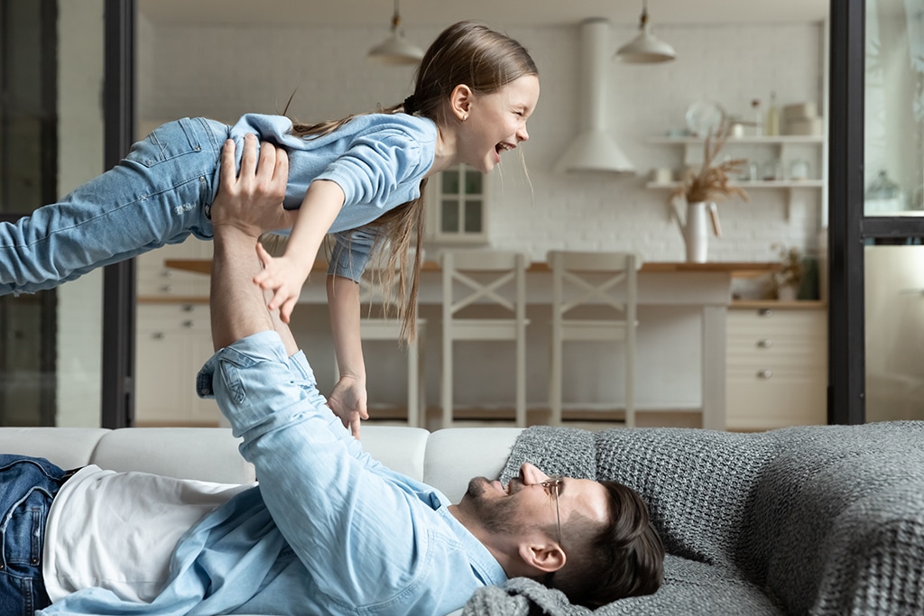 Loving young Caucasian father hold in hands lift up in air overjoyed little daughter, happy dad imitate plane have fun playing entertaining with excited small girl child in living room at home