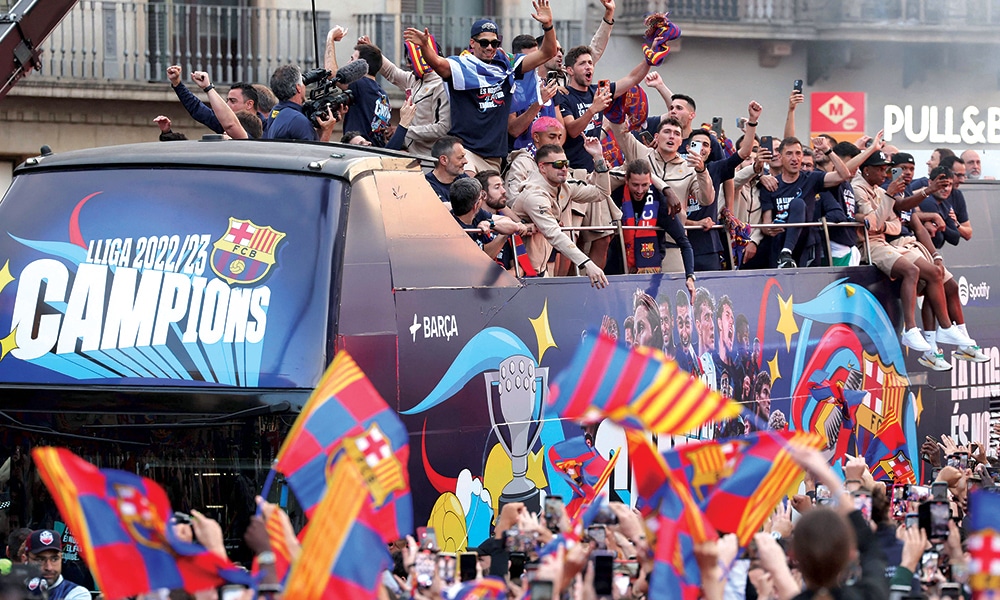 The players and staff of FC Barcelona men football team parade aboard a open-top bus followed by the women's team (out of frame), to celebrate their La Liga titles in Barcelona on May 15, 2023. The FC Barcelona women's team won its fourth consecutive La Liga title on April 30 after scoring 3-0 against Sporting de Huelva at the Estadi Johan Cruyff while the men's team yesterday won their 27th La Liga tile by thrashing Espanyol 4-2, wrestling the title back from rivals Real Madrid. (Photo by Lluis GENE / AFP)